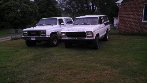 1979 Ford Bronco - A 1978 Ford Bronco and a GMC SUV are parked side-by-side on a grassy lawn next to a brick house.