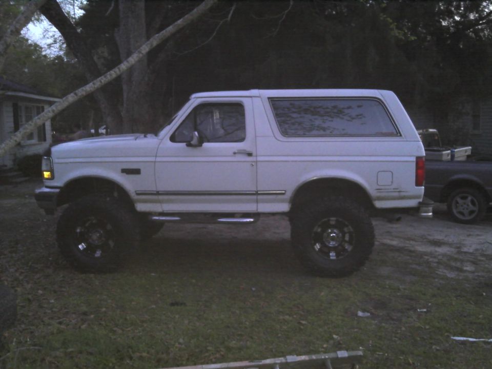 Lifted white SUV with aftermarket wheels parked on grass. Another dark truck is visible behind it