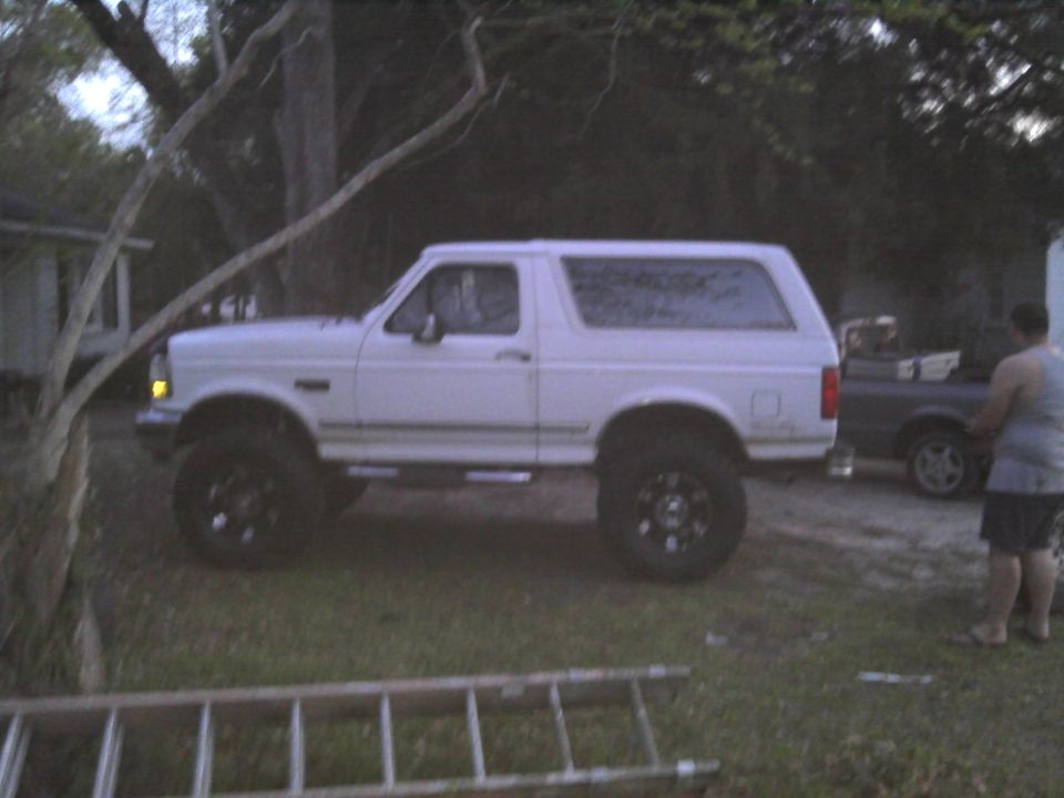 White Ford Bronco, lifted, with black wheels, parked on grass. A person stands near a dark colored car in the background