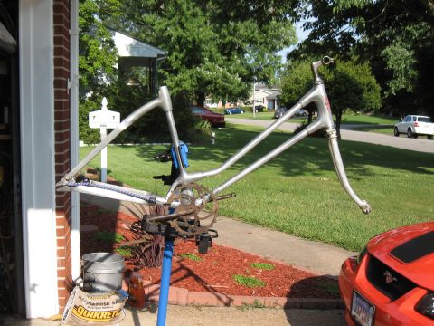 1994 Ford Bronco - A bicycle frame is mounted on a repair stand outdoors, with a residential street visible in the background and an orange car partially in f…