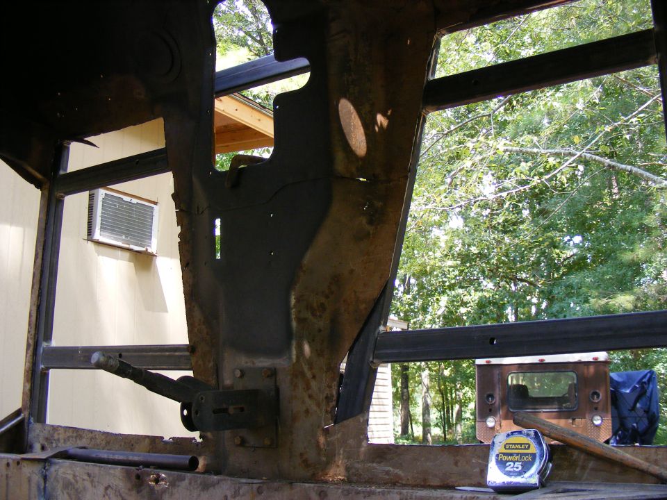 Inside of a vehicle's rusty frame, showing structural beams and a lever. Trees and a house are visible through openings