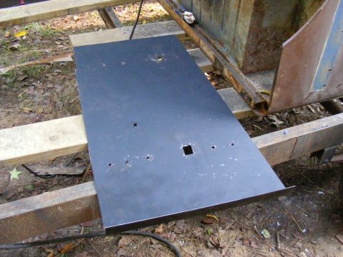 Flat black metal plate with holes and a square cutout, resting on wooden beams next to rusty metal framing