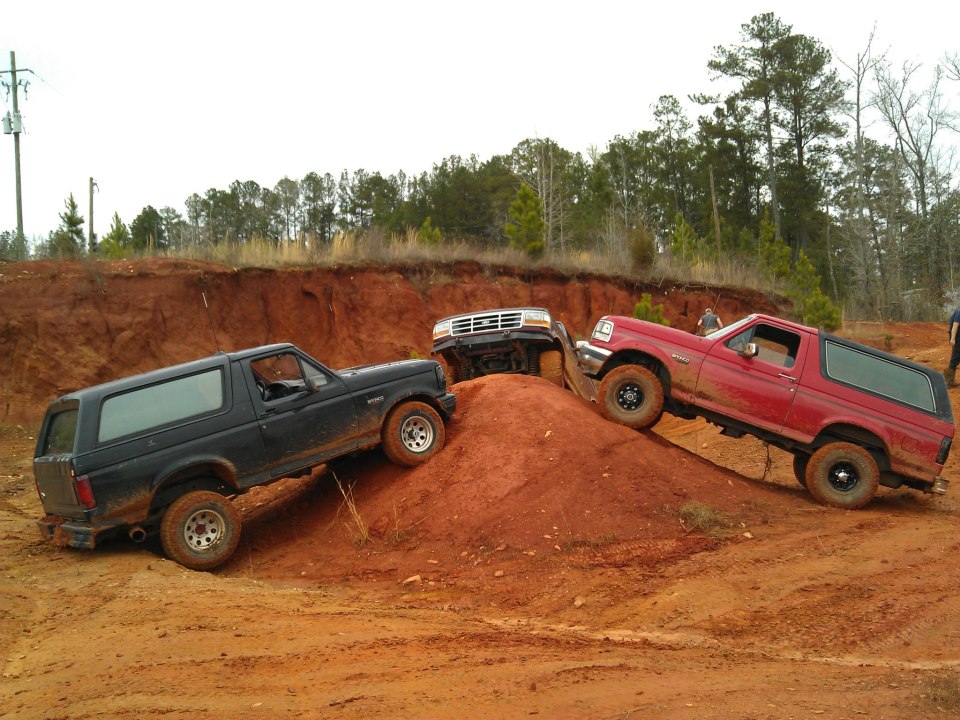 Three Ford Broncos climbing a dirt hill. One is black, one is red, and one is white