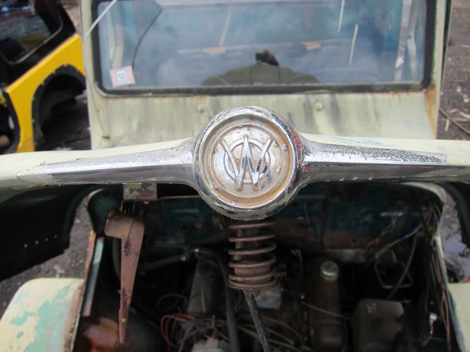 Close-up of a vintage jeep's front emblem with a 'W' inside a circle. Engine parts are visible below