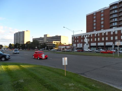 1971 Ford Ranchero - A bright red hot rod cruises down a city street, with several other vehicles and buildings visible.