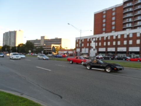 1971 Ford Ranchero - Several cars, including a black sports car and a red pickup, are driving on a street near buildings.