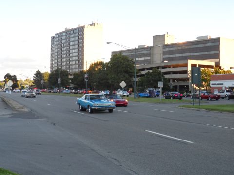 1971 Ford Ranchero - A blue 1969 Plymouth Road Runner coupe is seen driving on a road with other vehicles and buildings in the background.