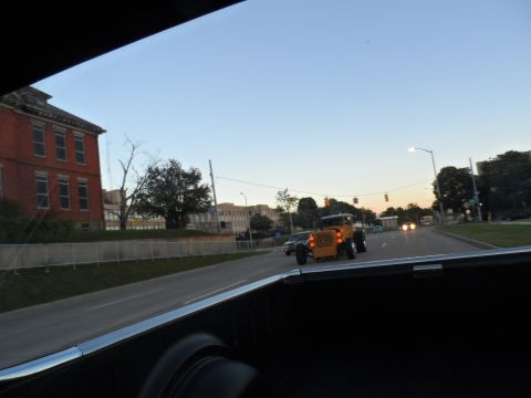 1971 Ford Ranchero - A yellow truck with prominent rear wheels is seen driving down a street at dusk, with other vehicles and buildings in the background.