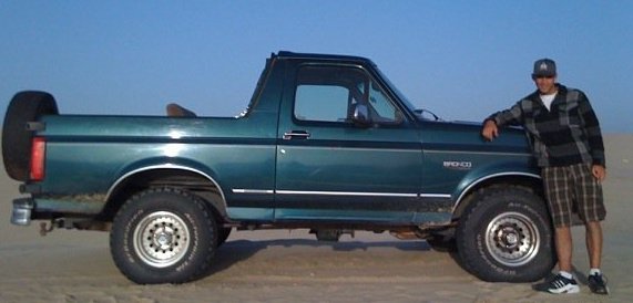 Green Ford Bronco with a spare tire on the back, parked on sand with a man standing next to it