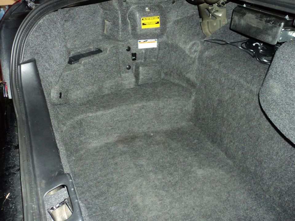 Car trunk interior with gray carpet, some wires visible, and a yellow warning sticker