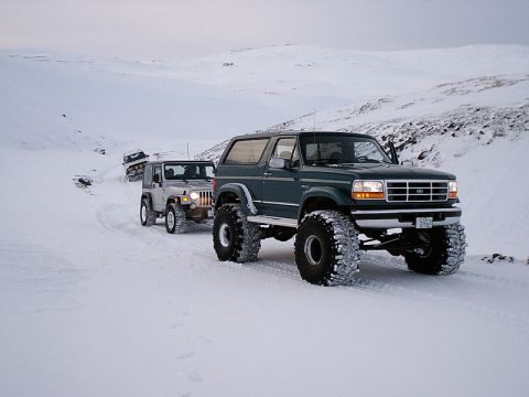1996 Ford Bronco - Several off-road vehicles navigate a snow-covered landscape, with a dark green Ford Bronco leading the pack.