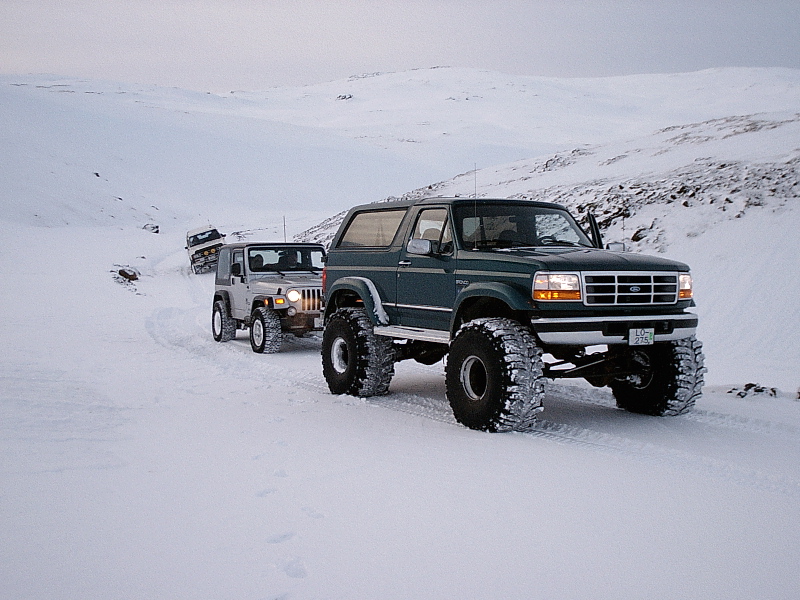 Lifted green Ford Bronco on big tires in snow, with a Jeep Wrangler behind on a snowy hillside.