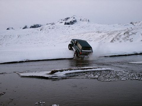 1996 Ford Bronco - 1996 Ford Bronco navigates a river during winter conditions with snow-covered mountains in the background.