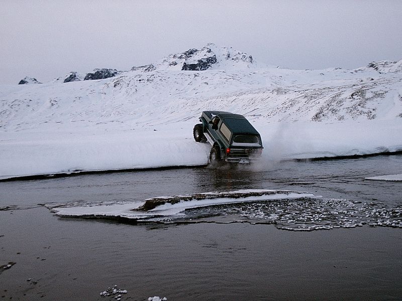 Dark green Bronco climbing snowy riverbank with front wheels lifted, spraying water