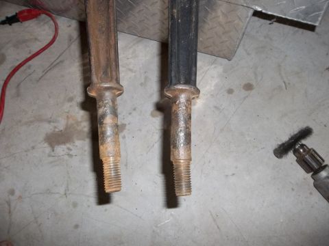 1995 Ford Bronco - Two metal shafts, one showing rust and the other painted black, sit on a concrete floor next to a wire brush tool.
