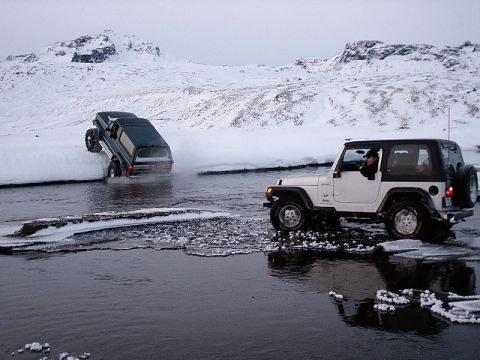1996 Ford Bronco - Two 4x4 vehicles navigate a cold, icy river with snow-covered mountains in the background.
