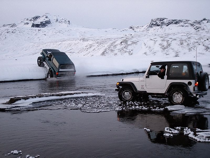 Green Bronco climbing snowbank and white Jeep Wrangler in icy river with snowy mountains