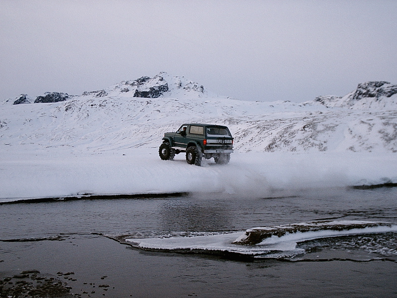 Lifted green Ford Bronco crossing a river in a snowy mountain landscape
