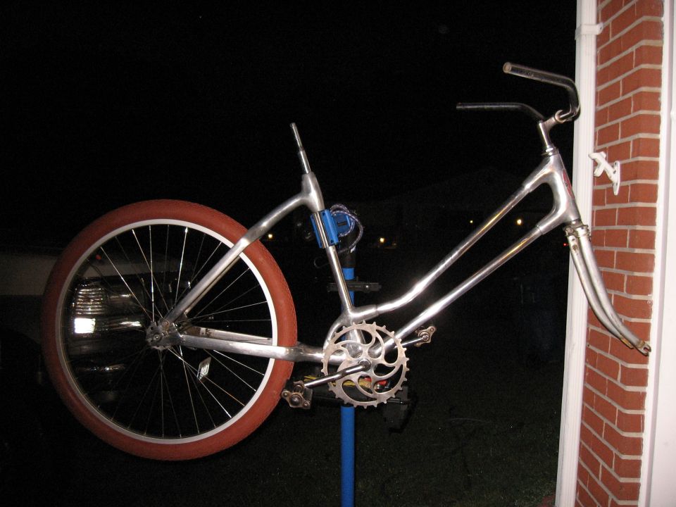 Chrome bicycle frame with a large red-orange tire on the rear wheel, leaning against a brick wall