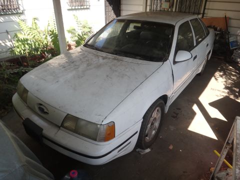 1990 Ford Taurus - A dusty white 1990 Ford Taurus sedan is parked under a carport, seen from the front three-quarter angle.