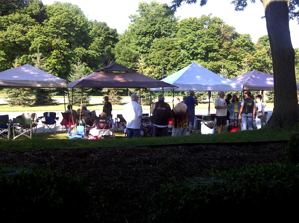 Outdoor event with several canopies set up, people gathered underneath and around them. Trees and grass in the background