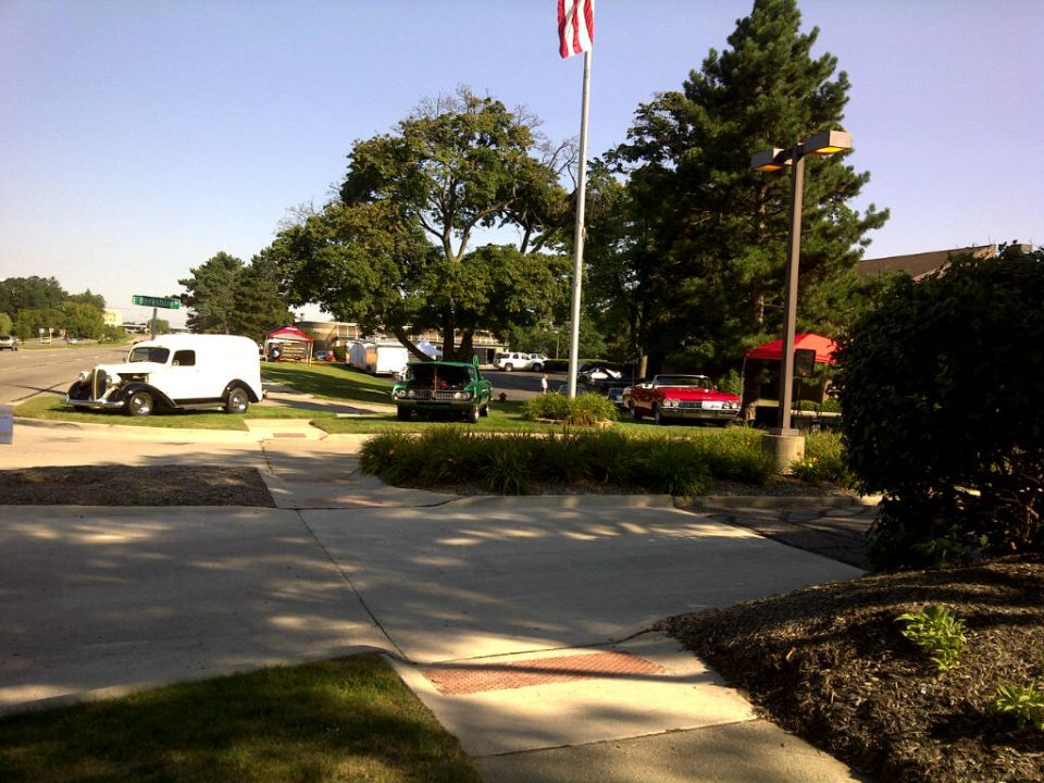 White panel van and a green pickup truck parked near a red convertible and a red kiosk under a flagpole