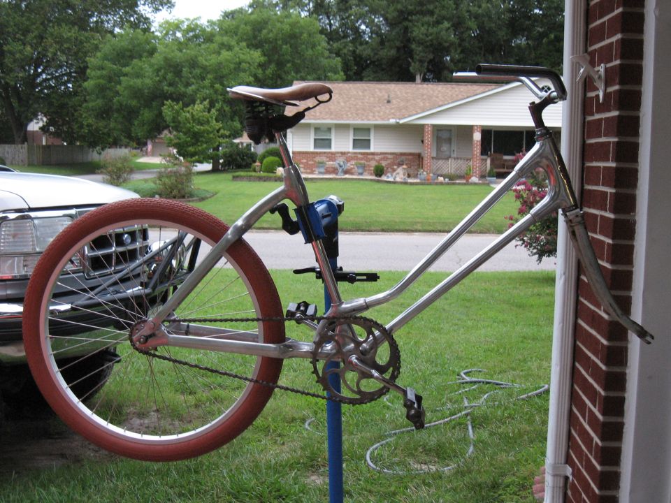 Chrome bicycle frame with a brown seat and red tire, mounted on a blue stand. Part of a silver truck is visible to the left