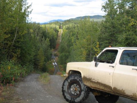 1989 Chevrolet C/K Pickup - A tan 1989 Chevrolet C/K Pickup navigates a muddy dirt road through a dense forest.