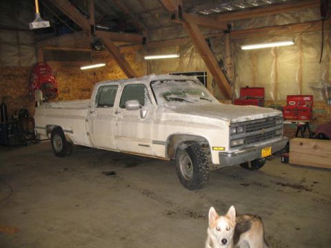 1989 Chevrolet C/K Pickup - A white pickup truck is parked inside a garage, partially covered in snow, with a dog looking at the camera.