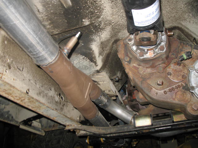 Underside view of a vehicle's exhaust pipes, driveshaft, and transfer case. A label is visible on a black component