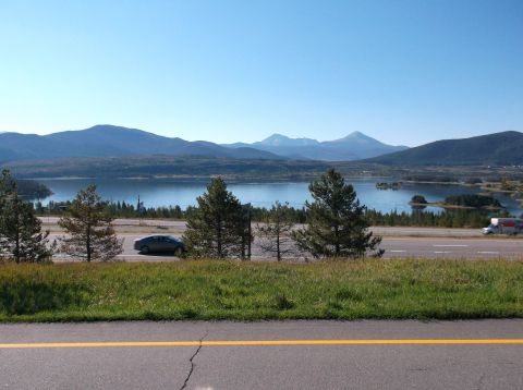 2009 Kenworth T800 - A car and a truck travel along a highway with a scenic lake and mountain backdrop under a clear blue sky.