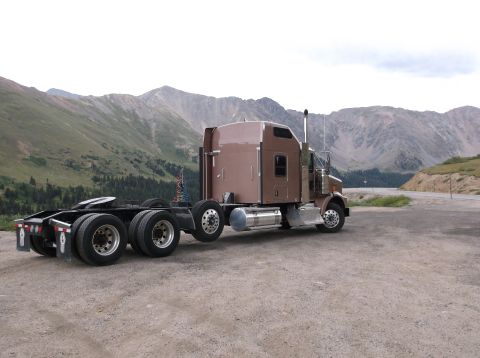 2009 Kenworth T800 - A brown semi-truck cab and chassis is parked on a gravel road with a mountainous landscape behind it.