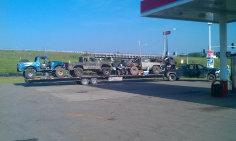 1987 Ford Bronco - A trailer carries multiple off-road vehicles, including trucks and ATVs, parked at a gas station under a bright blue sky.