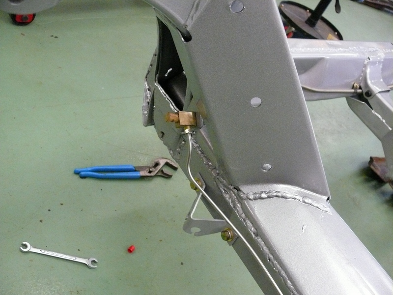 Close-up of a car's metal frame showing a brake line, a brass fitting, a wrench, and blue-handled pliers