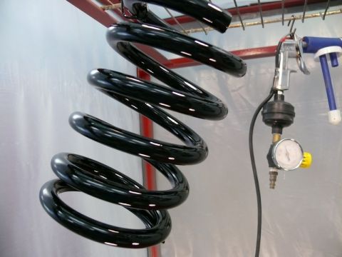1976 Chevrolet Corvette - A black coil spring is being processed with a spray gun and pressure gauge in a workshop.