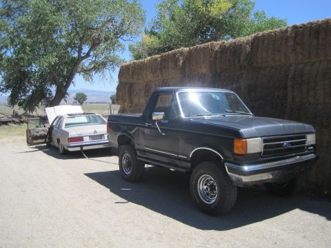 1989 Ford Bronco - A dark blue 1989 Ford Bronco is seen towing a beige sedan on a dirt road next to a large stack of hay bales.