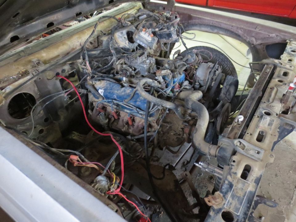 Disassembled engine bay with exposed engine, wiring, and parts. The vehicle appears to be in a state of disrepair or undergoing restoration