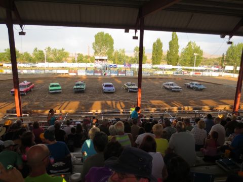 1989 Ford Bronco - Demolition derby cars gather on the dirt track under a covered grandstand.