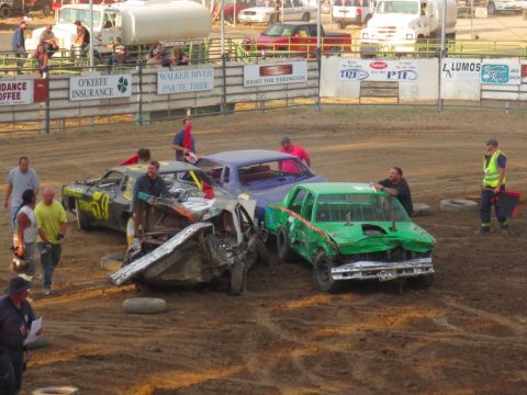 1989 Ford Bronco - Demolition derby cars sit in the dirt arena after a competition, with spectators and officials nearby.