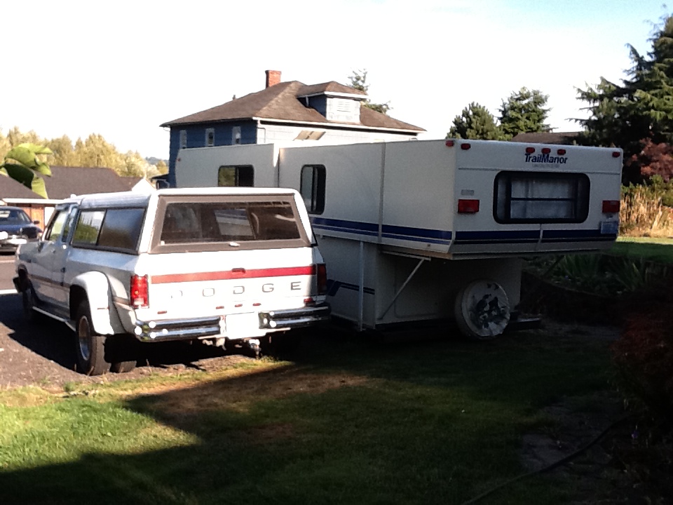 White Dodge pickup with a camper in tow. The truck has a cap and red stripe. The camper is white with blue stripes