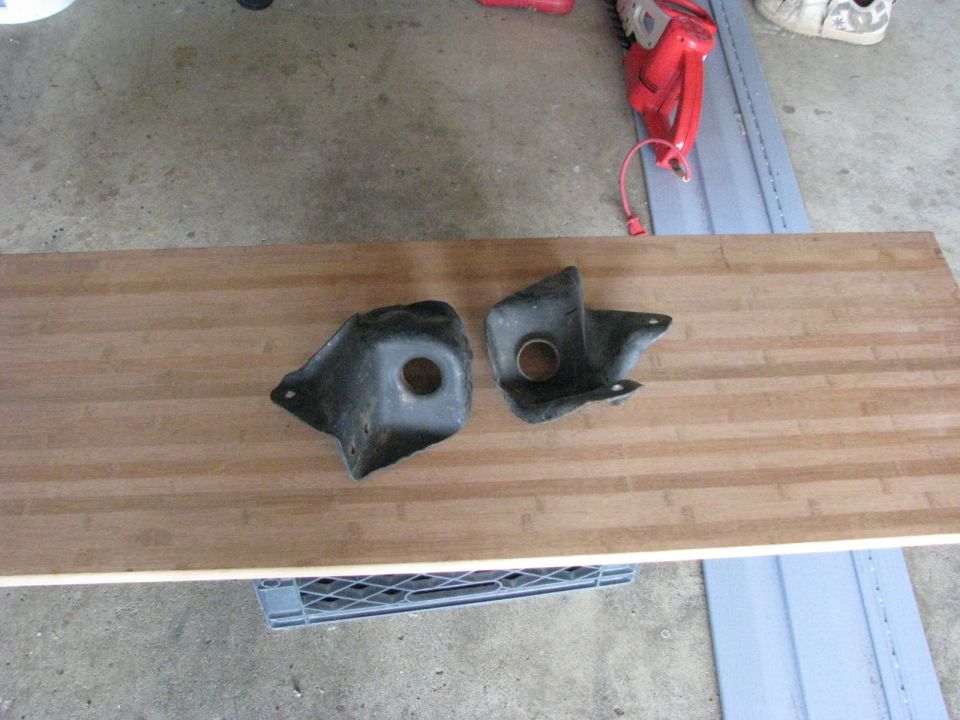 Two black metal brackets with holes, likely suspension components, laid out on a wooden surface
