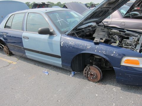 2001 Ford Crown Victoria - A two-tone 2001 Ford Crown Victoria sits in a junkyard with its hood up and missing wheels.