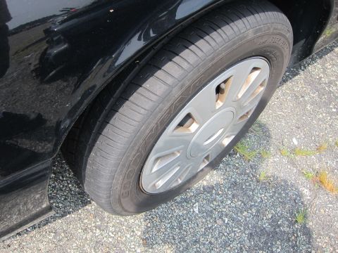 2001 Ford Crown Victoria - A close-up view of a black car's fender and wheel, showing the tire and hubcap against a gravelly surface.