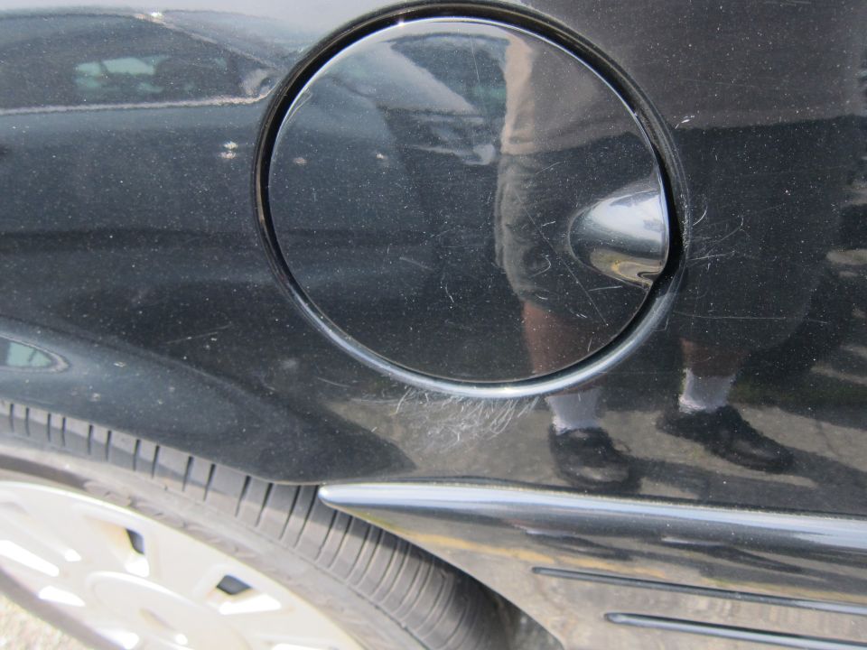Close-up of a black car's fuel door and rear quarter panel, showing scratches and a reflection of a person