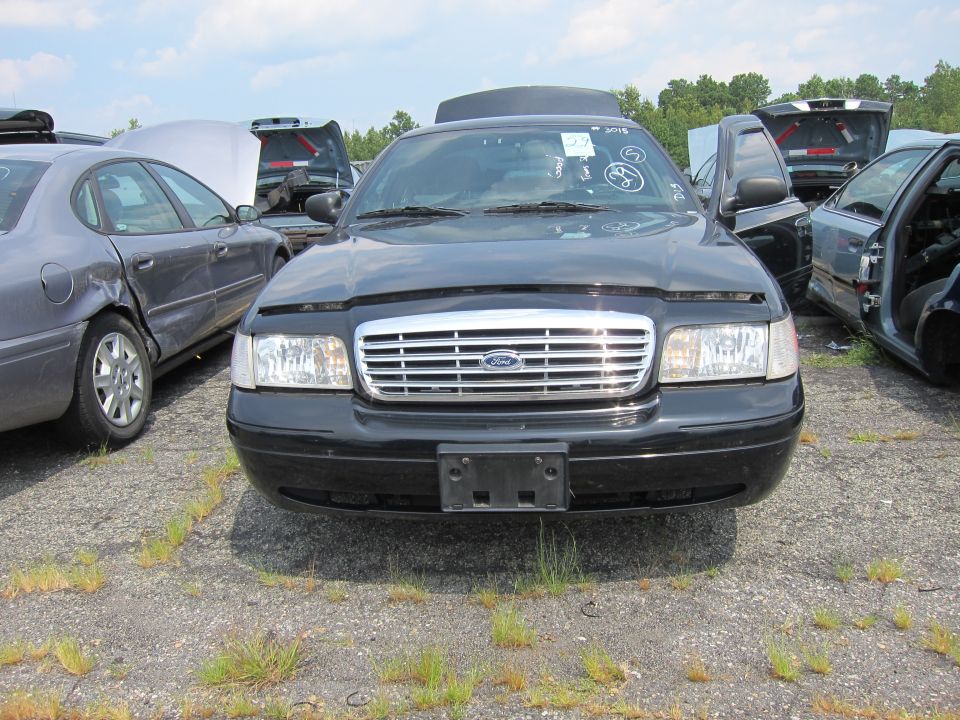 Front view of a black 2001 Ford Crown Victoria in a junkyard with other cars around