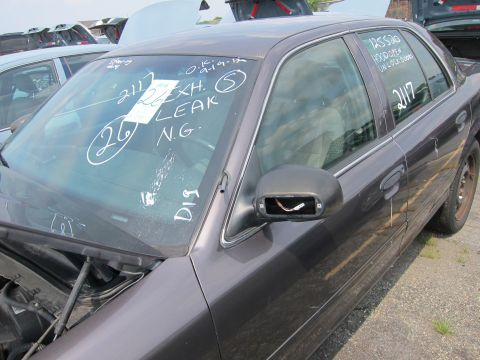 2001 Ford Crown Victoria - A dark sedan sits in a junkyard with notes on the windshield and a damaged side mirror.