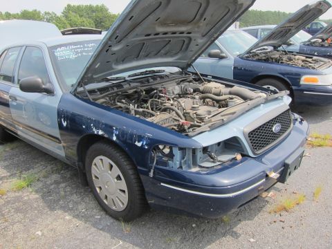 2001 Ford Crown Victoria - A blue and light blue Ford Crown Victoria sits in a junkyard with its hood open, revealing the engine compartment.