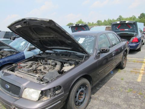2001 Ford Crown Victoria - A dark gray 2001 Ford Crown Victoria sits in a junkyard with its hood open, revealing the engine compartment.