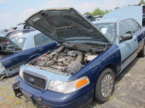 2001 Ford Crown Victoria - A blue and light blue 2001 Ford Crown Victoria sits in a junkyard with its hood open, revealing the engine compartment.