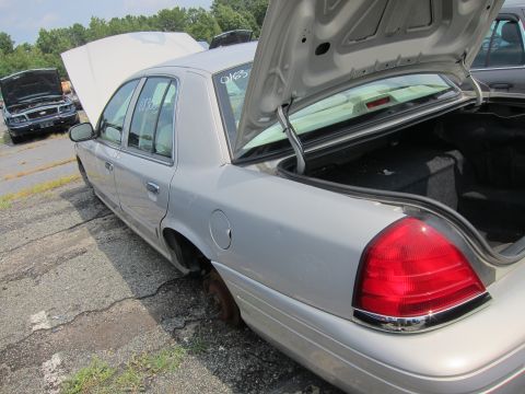 2001 Ford Crown Victoria - A silver 2001 Ford Crown Victoria sits in a junkyard with its trunk and hood open, missing a rear wheel.